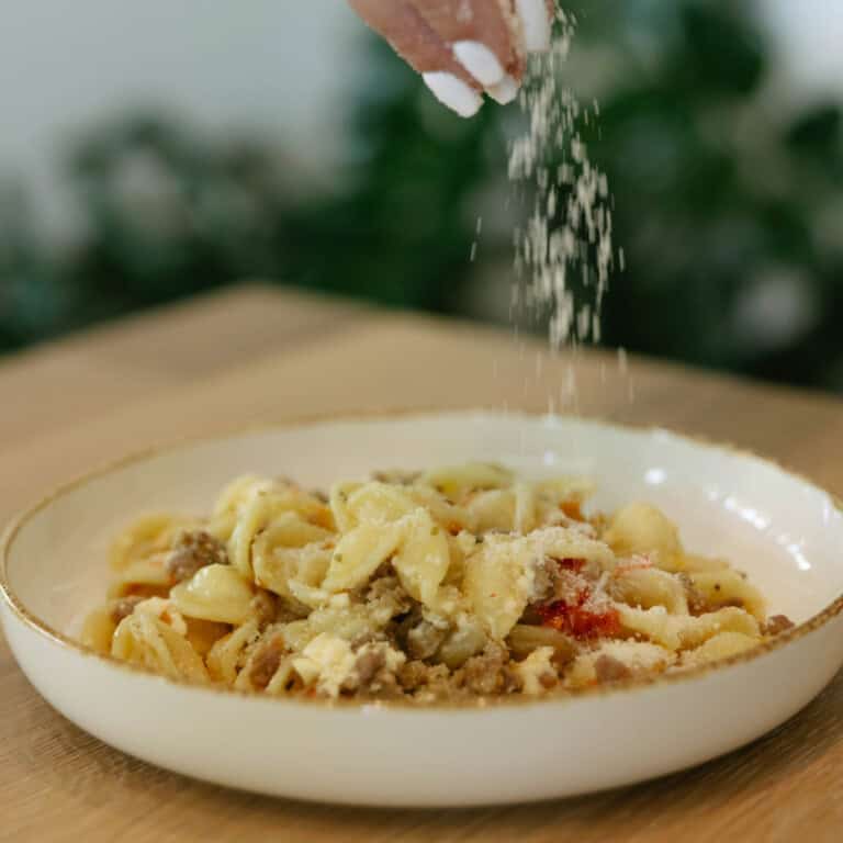 Parmesan being sprinkled on a bowl of creamy pasta with sausage peppers and feta.