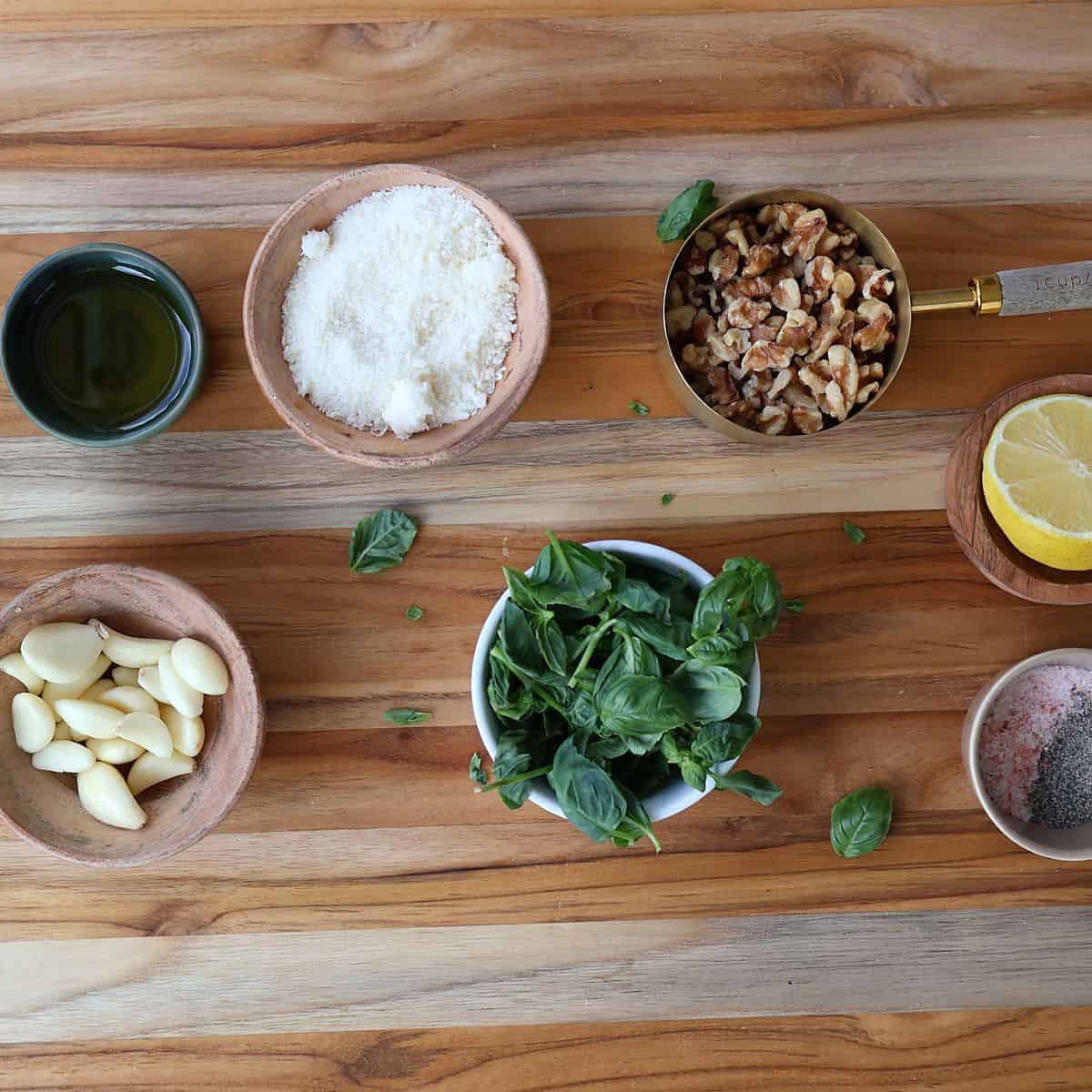 Ingredients for basil walnut pesto including walnuts, fresh basil, Parmesan cheese, garlic, olive oil, lemon, salt, and pepper in small bowls on a cutting board