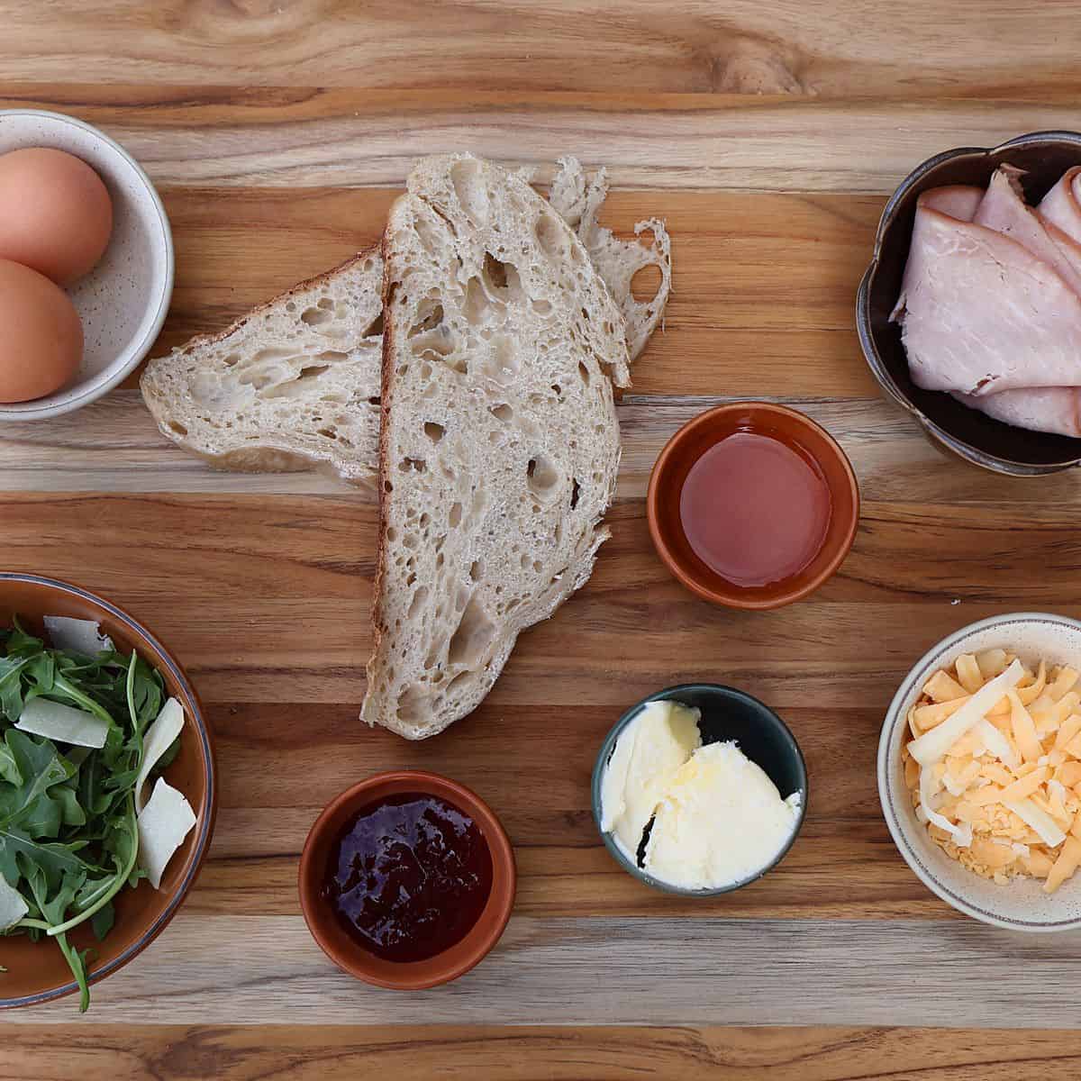 Ingredients for fried egg sandwich laid out on a wooden cutting board including eggs, ham, cheese, sourdough bread, arugula, and strawberry jelly