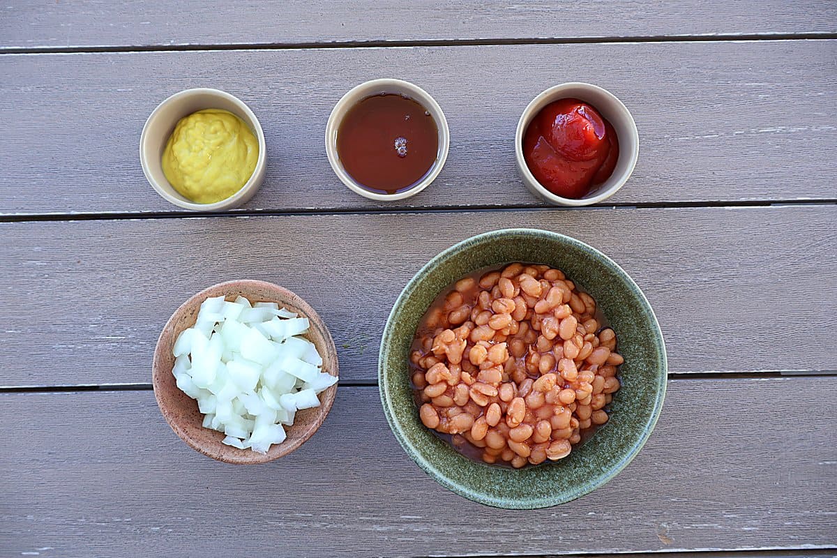 Ingredients for baked beans with canned beans laid out on a wood board