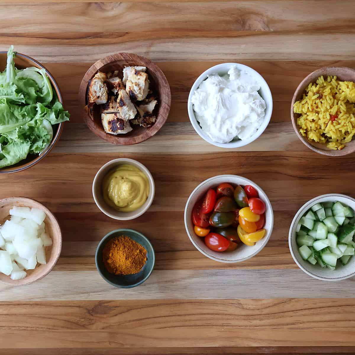 Ingredients for curry chicken bowl laid out including chicken breasts, Greek yogurt, yellow mustard, curry powder, rice, romaine lettuce, grape tomatoes, and red onion