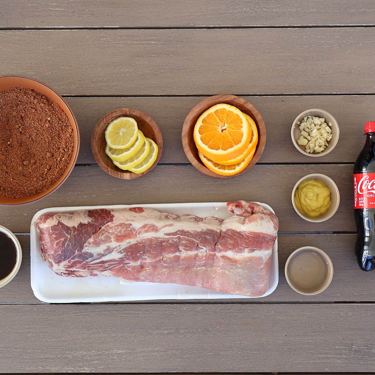 All ingredients for oven baked ribs recipe laid out including baby back ribs, Coca-Cola, coffee, brown sugar, spices, lemons, orange, and BBQ sauce