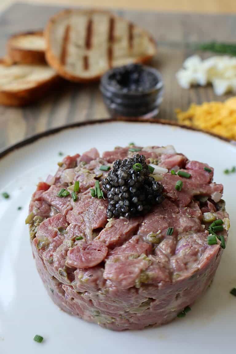Classic beef tartare on a white plate topped with caviar and chives, served alongside toasted baguette