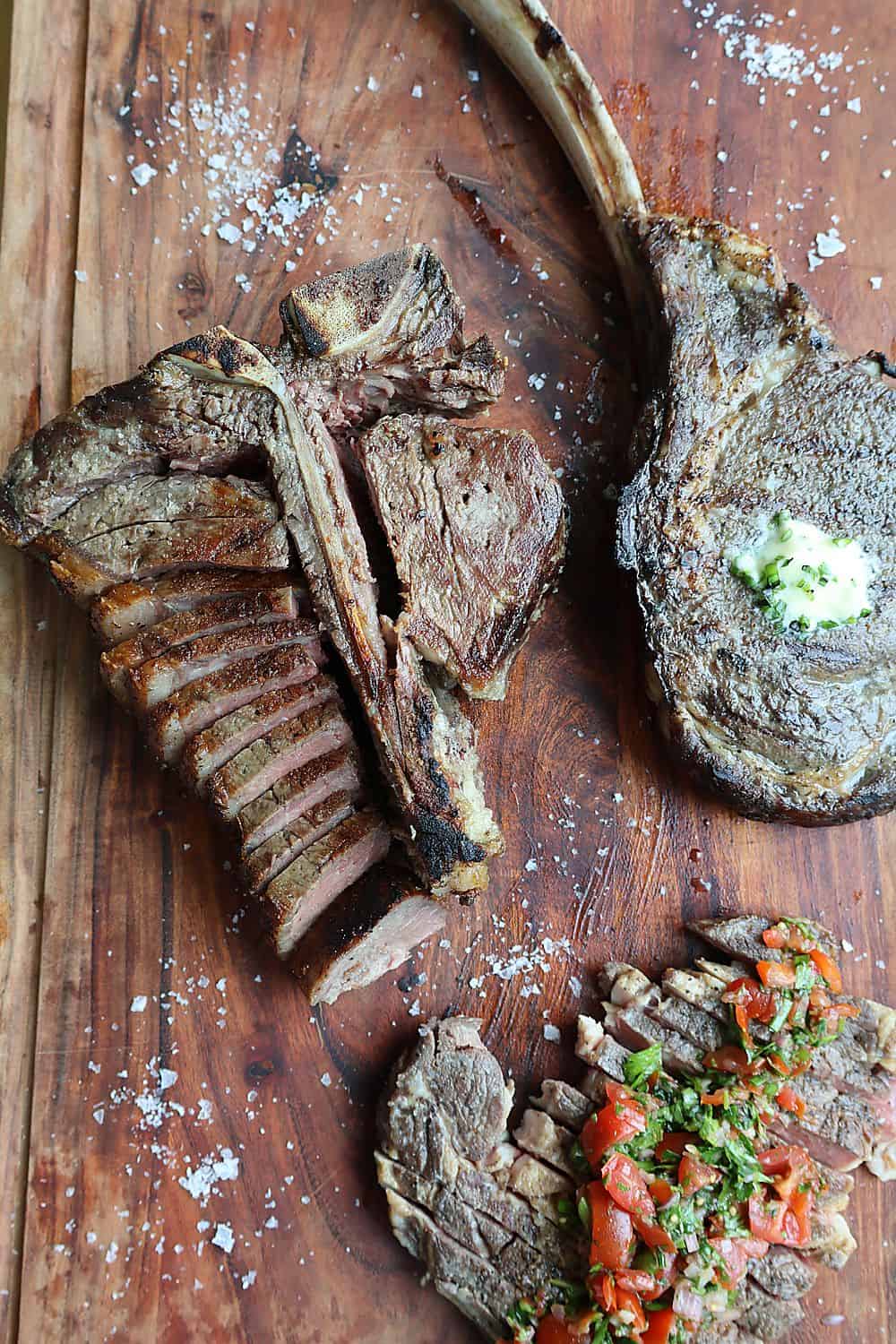 Sliced dry aged porterhouse steak on an end-grain wood board with the T-bone visible, garlic confit cloves, compound butter, and Florida Pure Sea Salt flakes