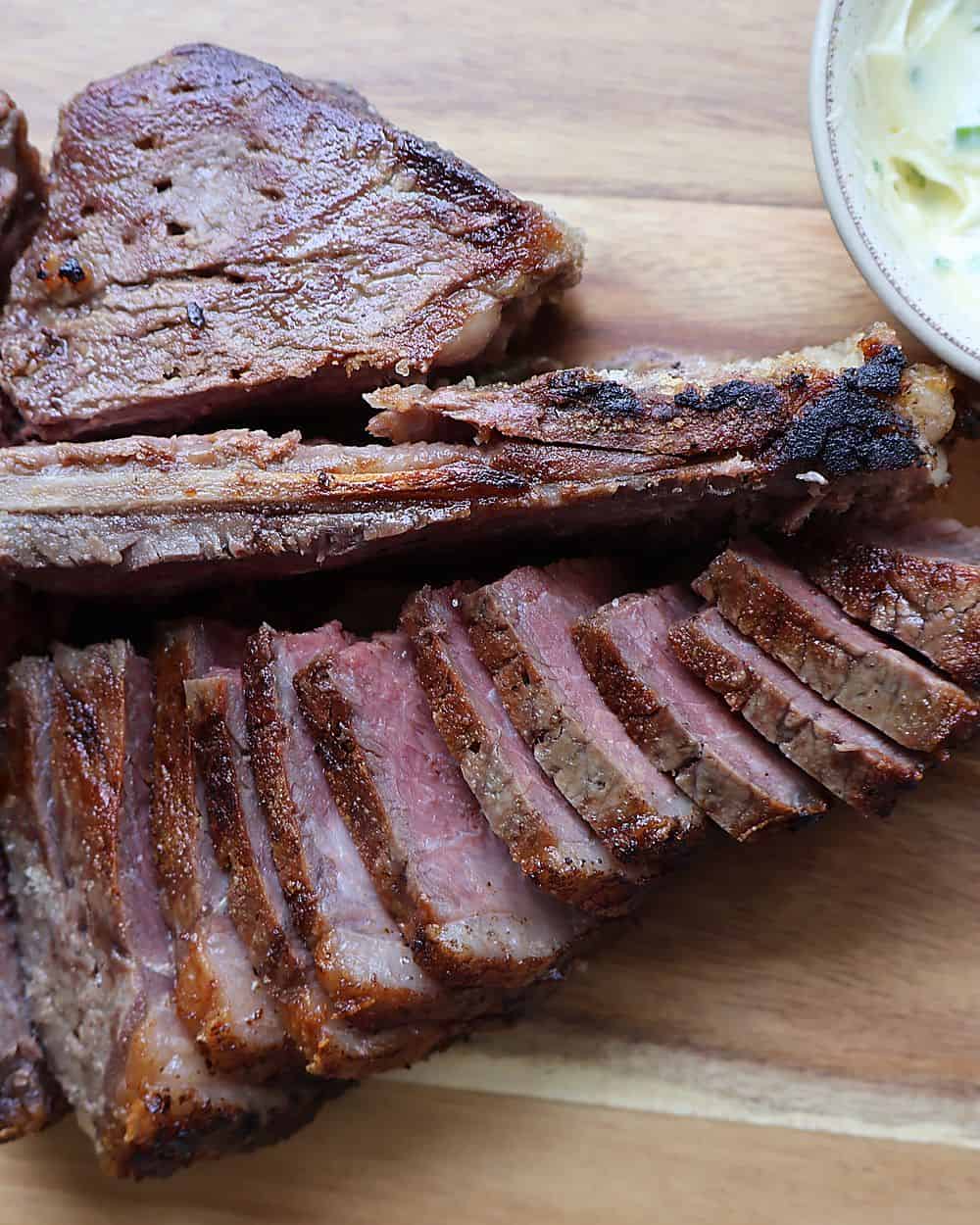 Sliced dry aged porterhouse off the bone on an end-grain cutting board with garlic confit cloves, compound butter, Florida Pure Sea Salt flakes, and a coarse grind of black pepper
