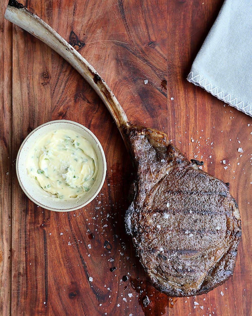 Seared reverse-sear tomahawk steak on a wood cutting board with grill marks visible, Cow's Rule compound butter in a small dish ready to be added