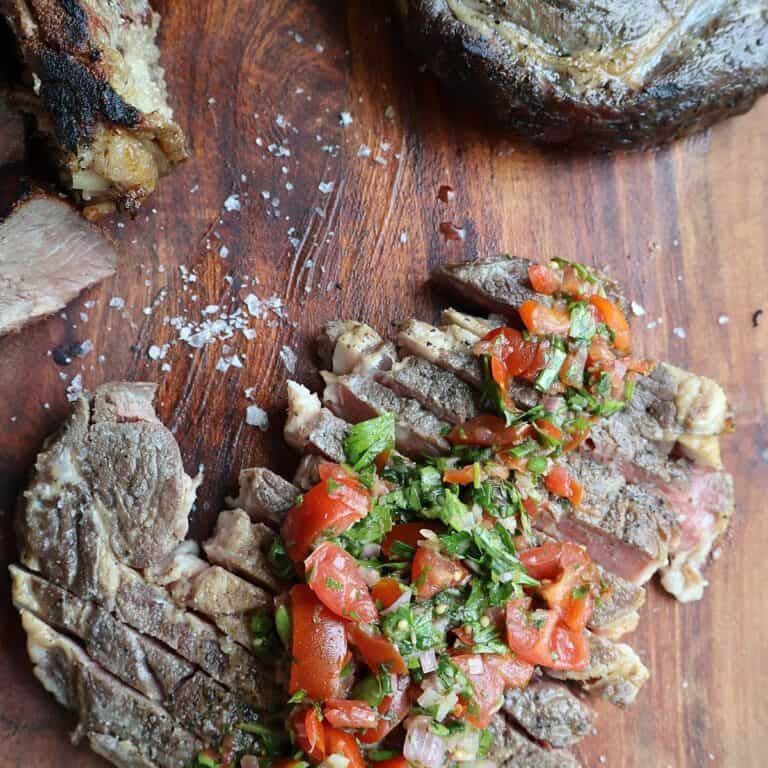 Sliced sous vide ribeye topped with tomato chimichurri and Maldon salt on a wooden board