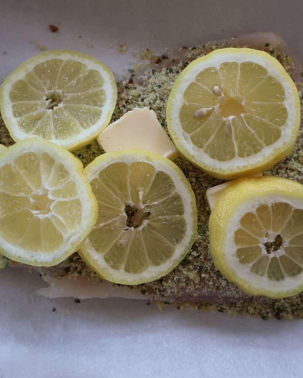 Crusted fish topped with butter pats and thin lemon slices before baking