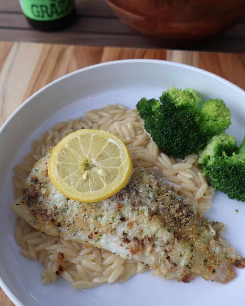 Served bowl of parmesan crusted fish over lemon orzo with broccoli and a lemon slice