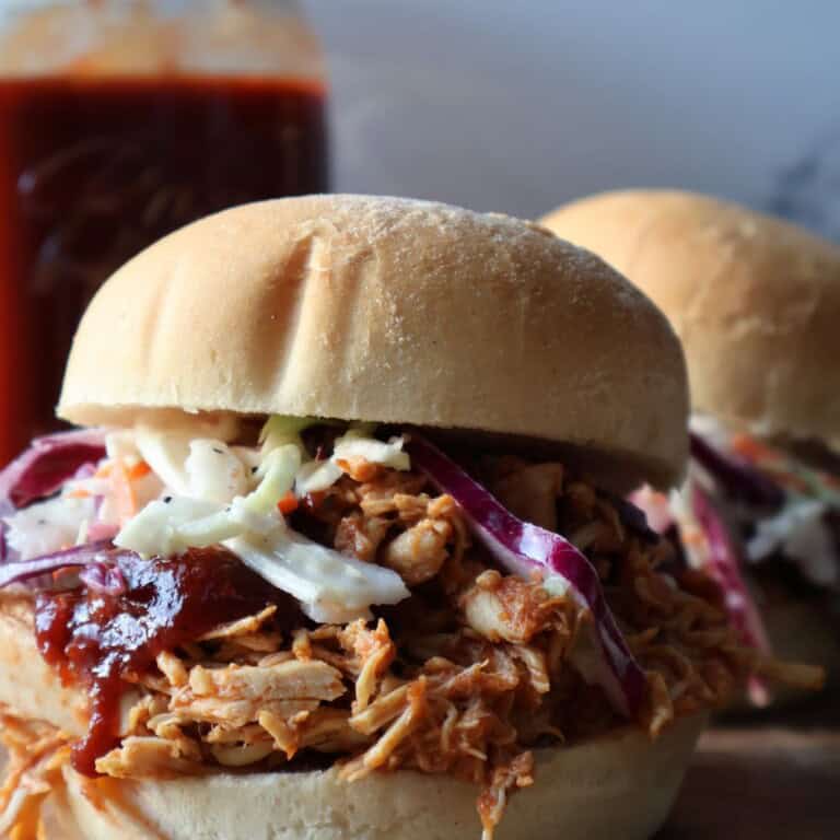 Two crock pot barbecue chicken sliders on soft buns topped with creamy coleslaw, a jar of homemade barbecue sauce behind them